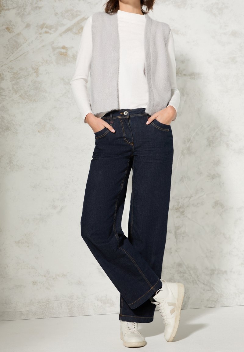 Femme portant un haut blanc à manches longues, un gilet sans manches gris clair, un jean large foncé et des baskets blanches, debout les mains dans les poches.