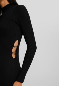 Abito nero a costine con maniche lunghe, caratterizzato da dettagli laterali a taglio accentuati da anelli metallici. Texture liscia e design aderente.