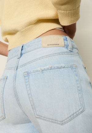 Jeans taille haute bleu clair avec poches arrière et étiquette blanche Stradivarius, portés avec un pull court jaune en tricot.