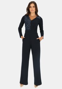 Granatowy kombinezon z długimi rękawami, teksturowaną kwiatową naszywką na górnej lewej stronie oraz bocznymi kieszeniami. Zwężane nogawki z gładkiego materiału.