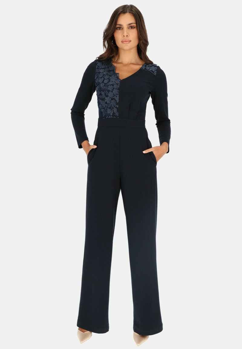 Granatowy kombinezon z długimi rękawami, teksturowaną kwiatową naszywką na górnej lewej stronie oraz bocznymi kieszeniami. Zwężane nogawki z gładkiego materiału.
