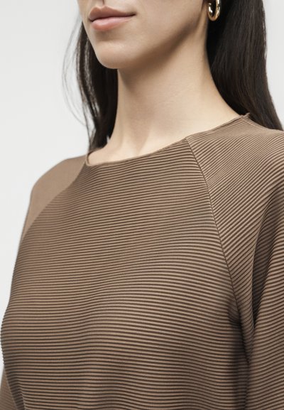 Γυναίκα που φορά χρυσό σκουλαρίκι κρίκο και καφέ ριμπ μπλουζάκι με λαιμόκοψη καραβοφάναρο, εμφανιζόμενη από το πηγούνι έως το πάνω μέρος του κορμού, με λευκό φόντο.