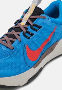 Chaussure de sport bleue avec une maille texturée et des matériaux synthétiques lisses. Présente un logo rouge proéminent, des lacets gris et une languette noire contrastante.