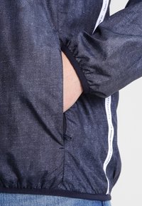 Marinblå jacka med strukturerad yta, med vita sidopaneler och elastiska ärmslut. Inkluderar en sidoficka för förvaring och elastisk dragsko.