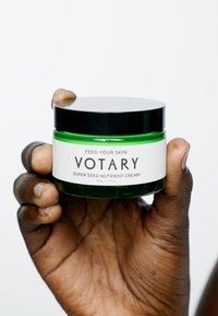 Grüner Glasbehälter mit schwarzem Deckel, beschriftet mit "Votary Super Seed Nutrient Cream." Das weiße Etikett trägt den Text "Feed Your Skin." Inhalt 50 ml.