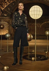 Blusa nera trasparente con motivi celestiali dorati, abbinata a pantaloni corti neri fino al ginocchio e stivali neri con tacco alto, in un ambiente decorativo.