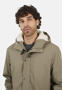 Veste imperméable vert olive avec une capuche doublée en fourrure, fermetures pression à l'avant et cordons de serrage. Texture lisse, design décontracté et coutures visibles.