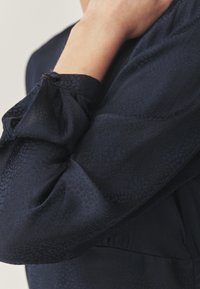 Blusa azul marino con tejido texturizado que presenta un sutil patrón de escamas. Mangas abullonadas con detalles fruncidos en los puños. Acabado suave en general.