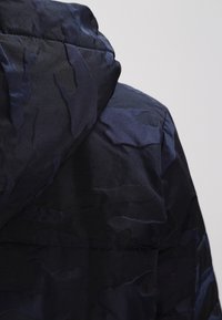 Chaqueta acolchada de color azul marino con un patrón de camuflaje texturizado y tonal. Cuenta con una capucha ajustable y un material liso y brillante.