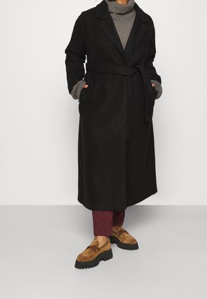 Persoon die een lange zwarte mantel met riem draagt over een grijze coltrui, bordeauxrode broek en bruine loafers met dikke zolen, staand met handen in de zakken.