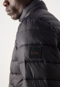 Zwarte pufferjack met gestikte secties, glanzende textuur, hoge kraag en een vierkant label met het "BOSS" logo.