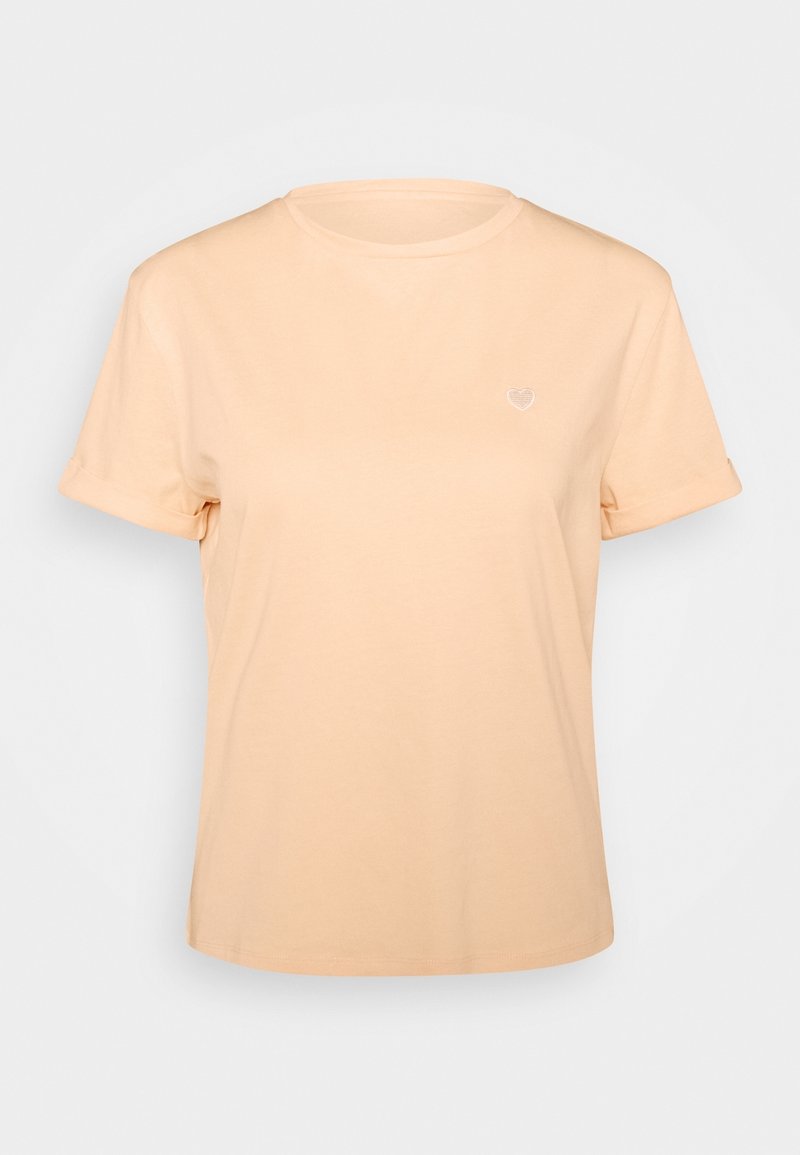 T-shirt à manches courtes couleur pêche clair avec revers roulés et un petit motif de cœur blanc sur la poitrine gauche, présenté sur un fond uni.