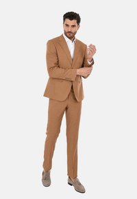 Completo marrone chiaro realizzato in tessuto texturizzato, caratterizzato da una giacca a due bottoni, pantaloni slim-fit e mocassini grigi. Camicia bianca sotto.