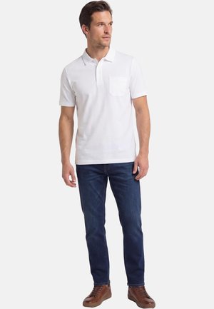 Mann trägt ein weißes Polohemd mit kurzen Ärmeln, dunkelblaue Jeans und braune Ledersneaker, steht vor einem einfarbigen Hintergrund.