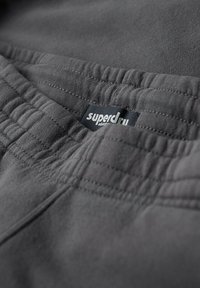 Grijze stof met een zachte textuur; heeft een elastische tailleband en een zichtbaar "superdry" label. Focus op de stikdetails.