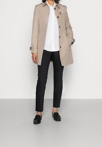 Person wearing beige trench coat with dark buttons, white shirt, black slim pants, and black loafers standing on gray floor.