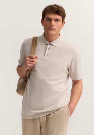 Junger Mann in beigem Polohemd und Hose trägt eine beiget Tasche über der Schulter, steht mit einer Hand in der Hosentasche vor einfachem Hintergrund.