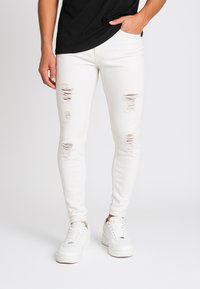 Valkoiset revenneet skinny-farkut, joissa on kuluneita yksityiskohtia, tiukka muotoilu ja sileä pinta. Yhdistetty valkoisiin lenkkareihin yksinkertaisella taustalla.