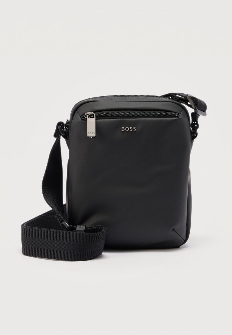 Συμπαγής μαύρη τσάντα ώμου με ρυθμιζόμενο ιμάντα, μπροστινή τσέπη με φερμουάρ και μεταλλικό λογότυπο "BOSS" σε λεία επιφάνεια.