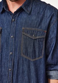 Denimhemd aus dunkelblauem Stoff, mit spitzem Kragen, Druckknöpfen und zwei aufgesetzten Taschen vorne mit kontrastierenden gelben Nähten.