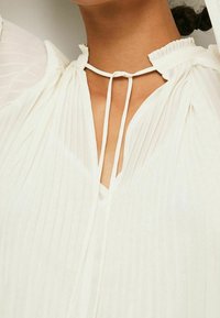 Vit plisserad blus med hög halsringning, fransad krage och ett knytband framtill, som visar upp ett lätt, genomskinligt tyg.