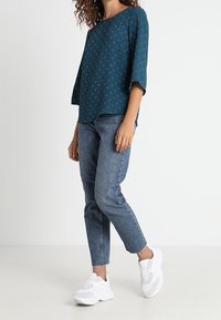 Mujer vistiendo una blusa de patrón en color verde azulado, jeans azules hasta el tobillo y zapatillas blancas, de pie contra un fondo sencillo.