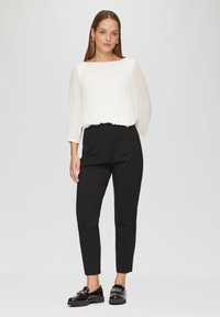 Witte geplooide blouse met lange mouwen, gecombineerd met zwarte hoge taillebroek en zwarte loafers met een glanzende afwerking. Achtergrond is eenvoudig wit.