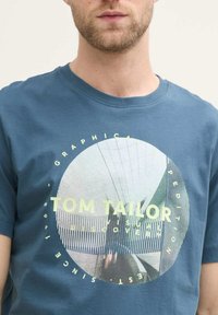 Sinine puuvillane T-särk, millel on ümmargune graafiline print, millel on helekollane tekst "TOM TAILOR" ja geomeetriline kujundus keskpunktis.