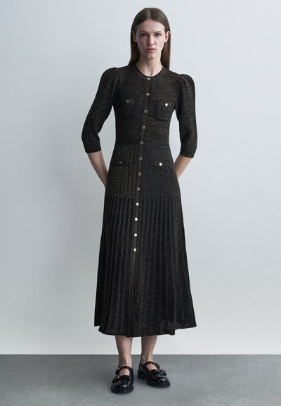 Femme portant une longue robe noire texturée avec des boutons sur le devant, des épaules bouffantes, des manches trois-quarts et des mocassins noirs plats, debout.