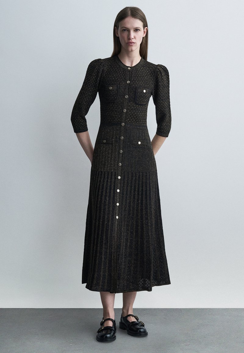 Γυναίκα που φοράει μακρύ μαύρο φόρεμα με υφή και κουμπιά στην μπροστινή πλευρά, με φουσκωτούς ώμους, μανίκια τριών τετάρτων και μαύρα επίπεδα μοκασίνια, όρθια.