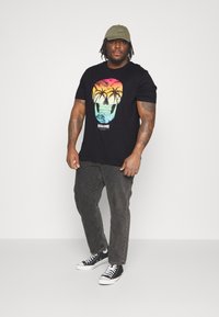 Black t-shirt with a gradient skull graphic in orange and turquoise, paired with gray jeans and black sneakers. Olive green cap on head.