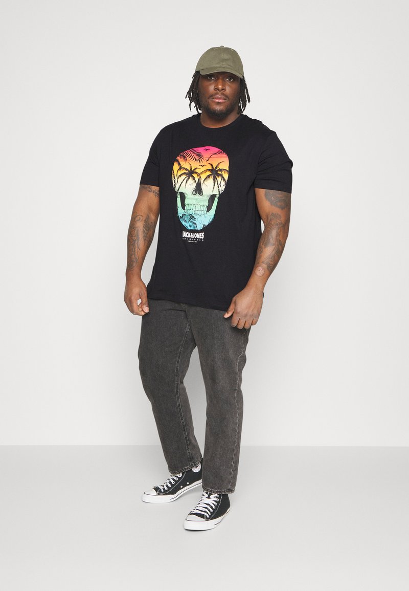 Black t-shirt with a gradient skull graphic in orange and turquoise, paired with gray jeans and black sneakers. Olive green cap on head.