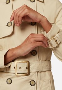 Krämfärgad lädertrenchcoat med dubbelknäppta knappar, bälte med guldfäste och upprullade manschetter. Detaljerad sömnad och stora matchande knappar.