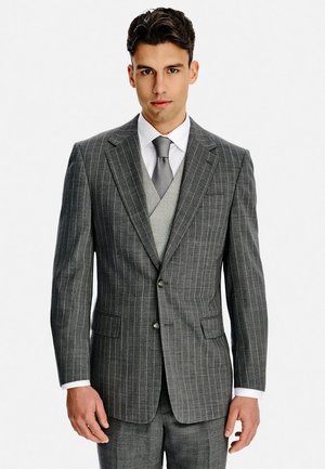 Mężczyzna ubrany w szary garnitur w prążki z dopasowaną kamizelką, białą koszulą i szarym krawatem, stojący na jednolitym białym tle.