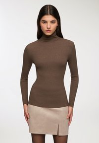 Maglione a collo alto a coste marrone con maniche lunghe, dal design aderente. Indossato con una mini gonna beige chiaro, con una sottile apertura frontale.