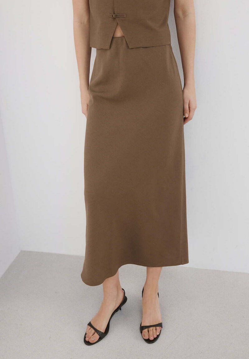 Femme portant une longue jupe marron et un gilet assorti, avec des sandales à talons noires à bout ouvert, debout contre un mur clair uni.