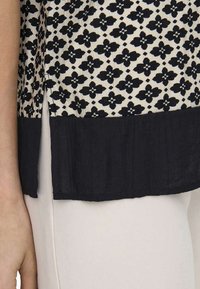 Blouse à motifs noir et blanc avec design floral. Présente un tissu rigide et un ourlet noir uni en bas. Détail d'ouverture sur le côté.