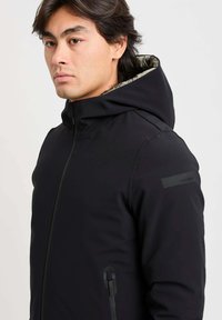 Giacca nera con cappuccio, finitura opaca, interno testurizzato, zip frontale e tasche laterali. Presenta un marchio discreto sulla spalla.