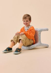 Bambino seduto su una sedia a macchie blu chiaro. Indossa un maglione arancione, una maglietta a righe, pantaloni color khaki e scarpe verdi con dettagli arancioni.