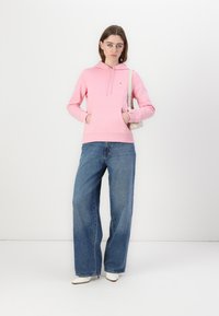 Haut à capuche rose clair avec une poche avant et un cordon de serrage, associé à un jean bleu à jambe large et des bottines blanches à talons. Design simple.
