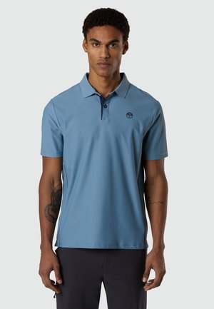 Homme aux cheveux courts et bouclés portant un polo bleu clair et un pantalon noir, debout devant un fond gris uni.