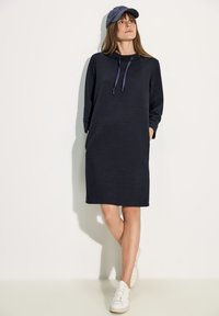 Marinblå oversized hoodieklänning med texturerad tyg, som har en dragsko i huvan och sidofickor, kombinerad med vita sneakers och en keps.