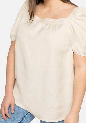 Femme portant un chemisier crème à manches courtes bouffantes et col carré, associé à un jean bleu.