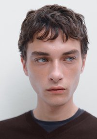 Männlicher Model mit kurzem, welligem braunem Haar und blauen Augen, der einen dunkelbraunen V-Ausschnitt-Pullover über einem marineblauen Hemd trägt, vor einem hellen Hintergrund.