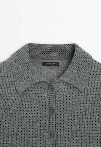 Polo en maille gris avec de petits trous carrés, col texturé et boutons foncés sur le devant, posé à plat sur fond blanc.