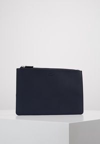 Pochette rectangulaire bleu marine avec fermeture éclair, surface texturée et petit logo crocodile embossé centré près du bord supérieur.