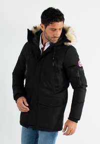 Parka noir avec capuche en fausse fourrure, zips et deux poches avant. Présente des poignets côtelés et un écusson logo sur le bras.