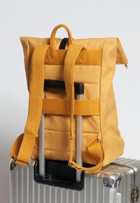 Zaino in tessuto giallo con design imbottito, spallacci regolabili e manico superiore, posizionato su una valigia argentata con ruote.