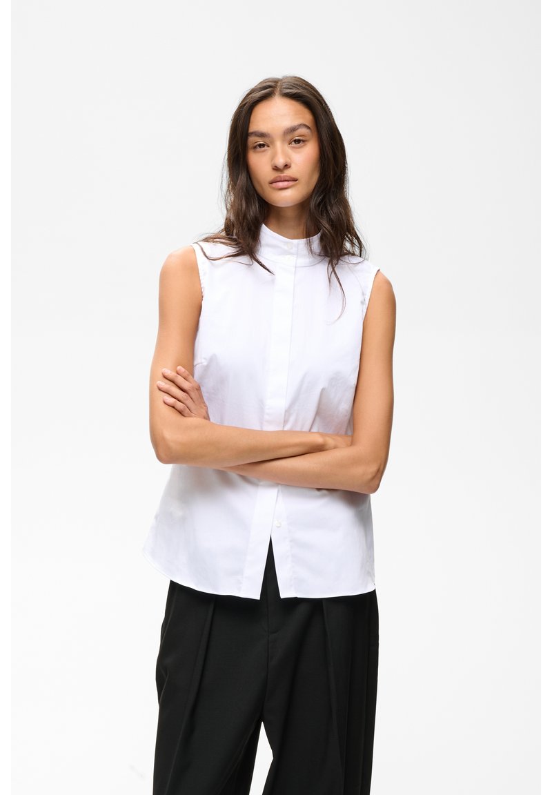 Femme aux longs cheveux foncés portant un chemisier blanc sans manches et un pantalon noir, debout les bras croisés sur fond blanc.