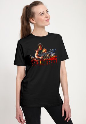 Jeune femme avec queue de cheval brune portant un t-shirt noir avec Rambo tenant une grande arme avec des flammes rouges derrière.